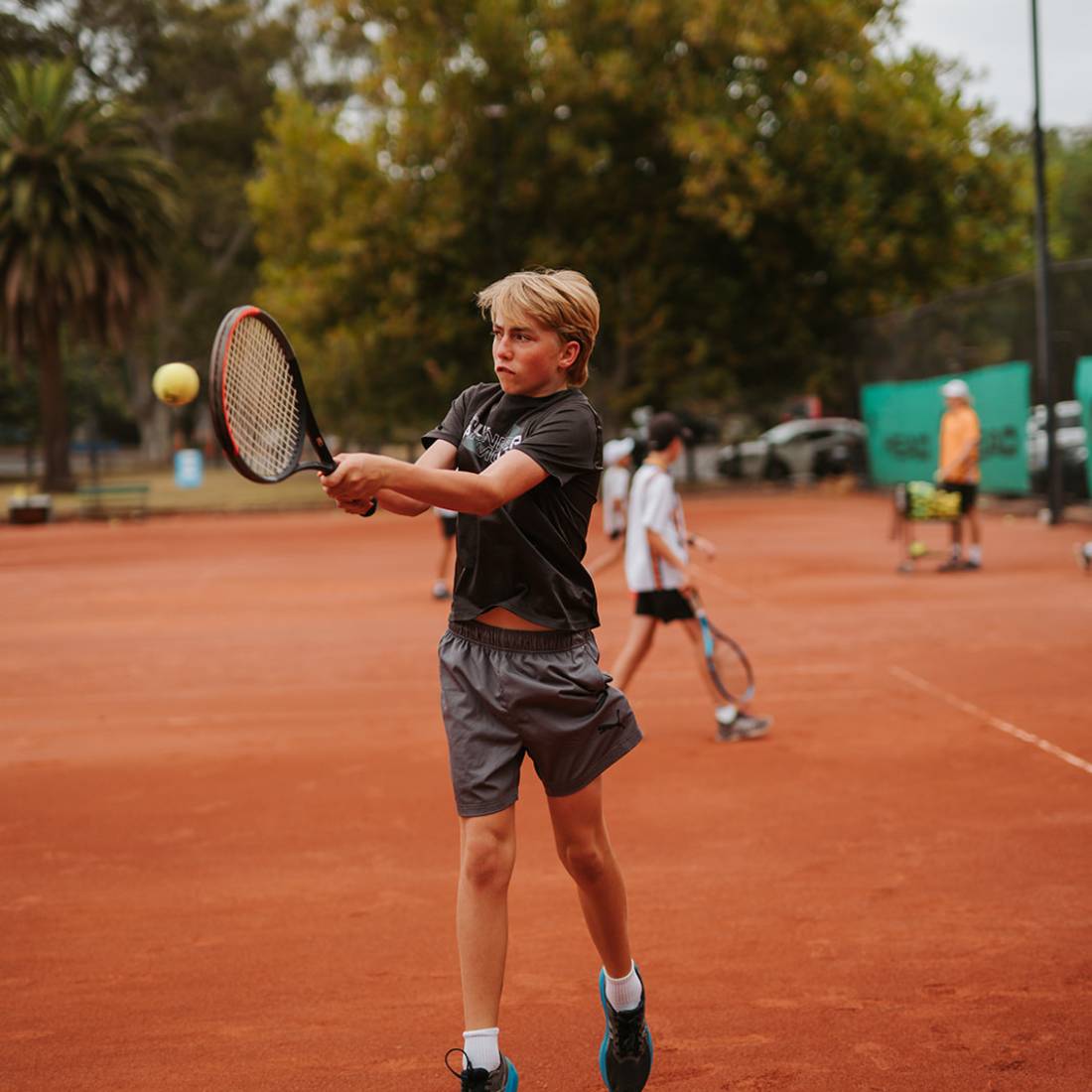 Young boy playing tennis on an outdoor clay court, hitting a ball with a backhand swing.