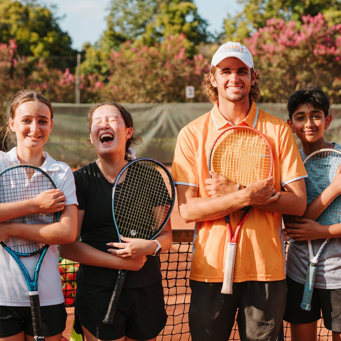 Four smiling young tennis players holding rackets, standing on a tennis court with greenery in the background.